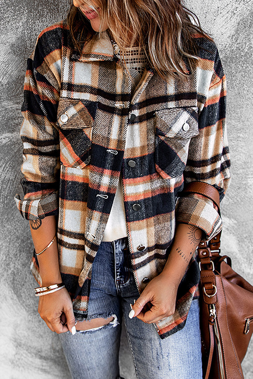 A person wearing a Brown Plaid Button Up Long Sleeve Flannel Shacket layered over a white top with ripped jeans and holding a brown handbag stands against a textured gray wall.