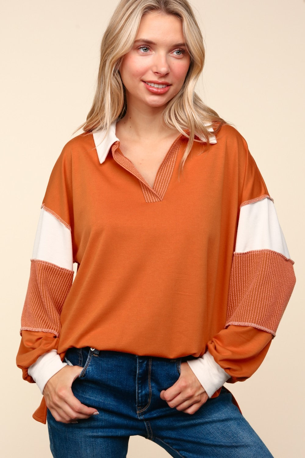 A person with blonde hair is wearing the Haptics Color Block Exposed Seam Long Sleeve Top in orange with white and orange sleeves, paired with blue jeans, standing against a beige background.