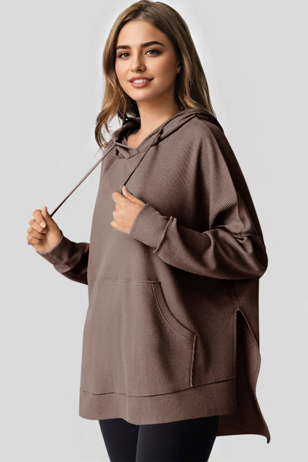A woman with long hair is standing in profile, wearing a Coffee Waffle Knit Fleece Lined High Low Oversized Hoodie and black shorts. The background is a plain light gray.