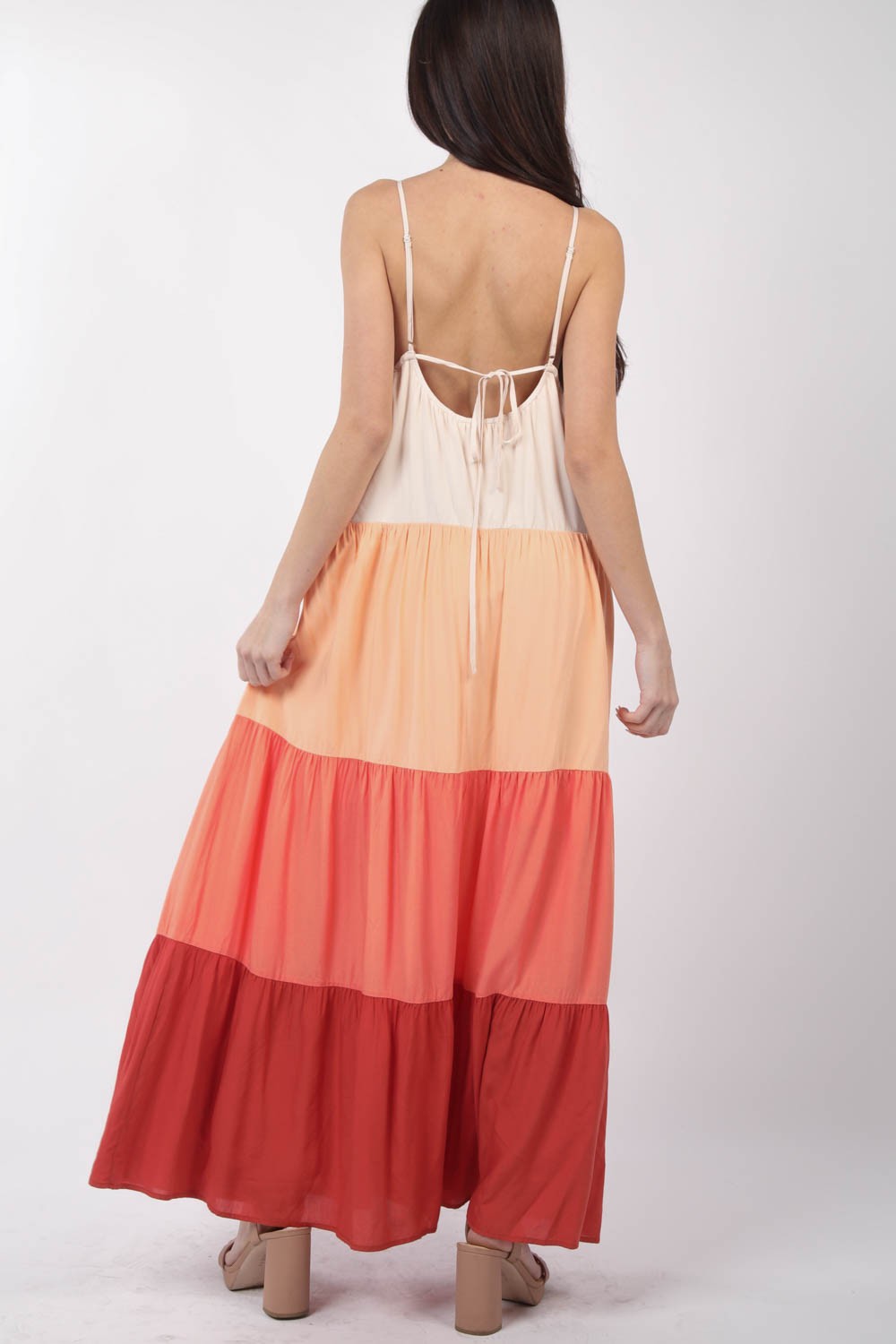 A woman stands, wearing the VERY J Color Block Tiered Maxi Cami Dress in shades of beige, peach, and red, paired with beige sandals. Her right hand holds the side of her dress as she poses against a plain white background—a truly fashionable choice.