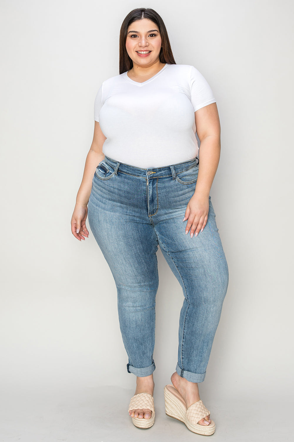 A person wearing a white shirt, Judy Blue Full Size Cuffed Hem Slim Jeans, and black wedge-heeled sandals is standing in a room with beige chairs and plants in the background.