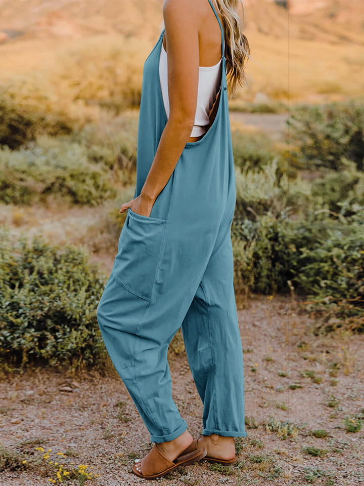 Person wearing a Double Take Full Size V-Neck Sleeveless Jumpsuit with Pockets and sandals, walking outdoors on a dirt trail with bushes in the background.