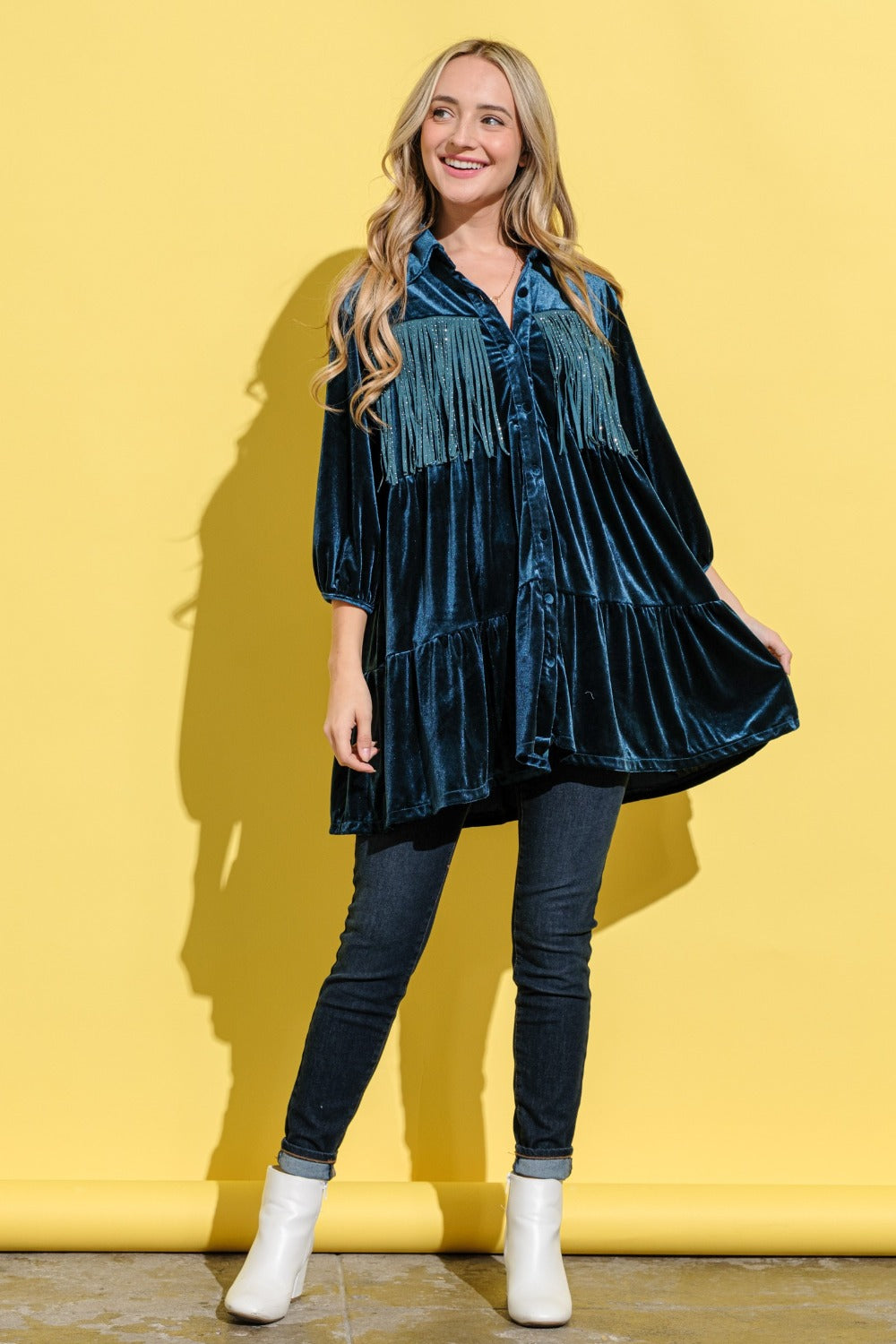 A woman in the And The Why Fringe Detailed Velvet Shirt Dress with a tailored fit and button-up front, wearing jeans, stands against a yellow background.