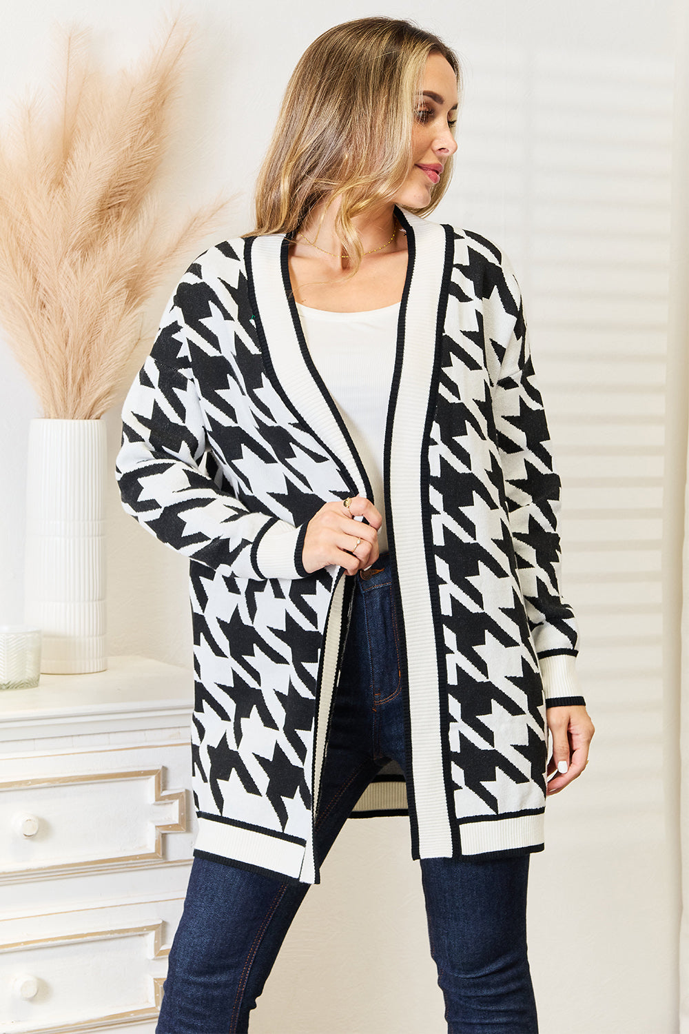 Woman wearing the Mandy Woven Right Houndstooth Open Front Longline Cardigan over a white top, standing next to a white chest of drawers with pampas grass in a vase on it.