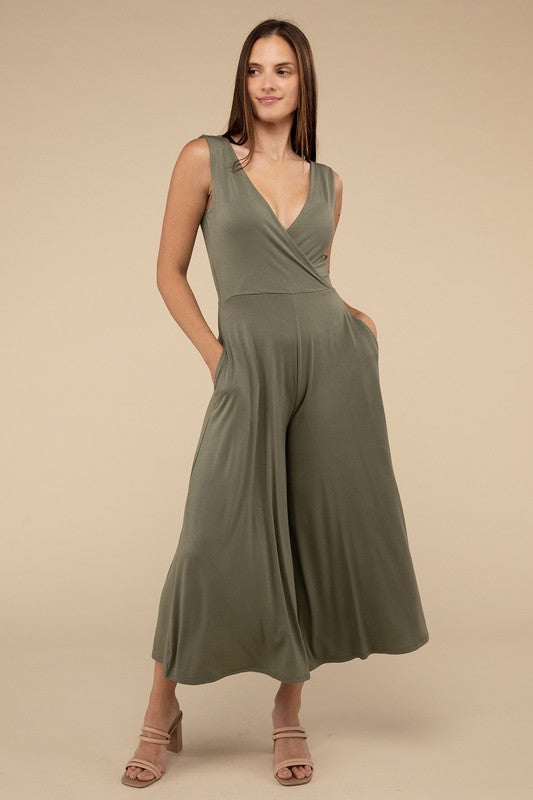 A woman stands in a studio, wearing an elegant olive green Surplice Neckline Sleeveless Jumpsuit with wide legs and paired with beige heels.
