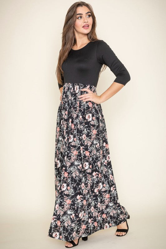 A woman elegantly stands in a room with wooden flooring and modern furniture, wearing a ditsy floral maxi dress and a long-sleeve navy top.