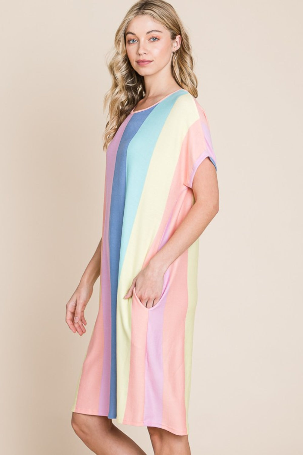 A person stands against a plain background, wearing the BOMBOM Striped Short Sleeve Dress with Pockets, crafted from comfortable fabric and featuring pastel stripes, with hands casually tucked into the convenient pockets.