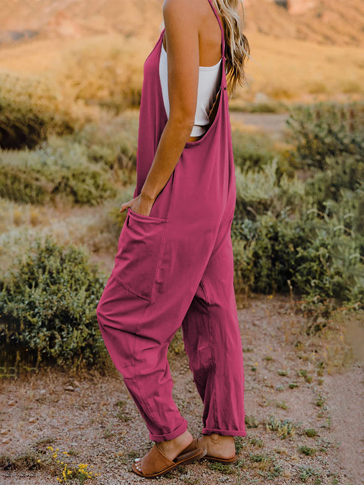 A woman stands outdoors wearing the Double Take Full Size Sleeveless V-Neck Pocketed Jumpsuit in a loose-fitting, imported brown design over a white top. Her surroundings include grass, bushes, and a rocky landscape in the background. The jumpsuit's polyester fabric ensures comfort and durability with every machine wash.