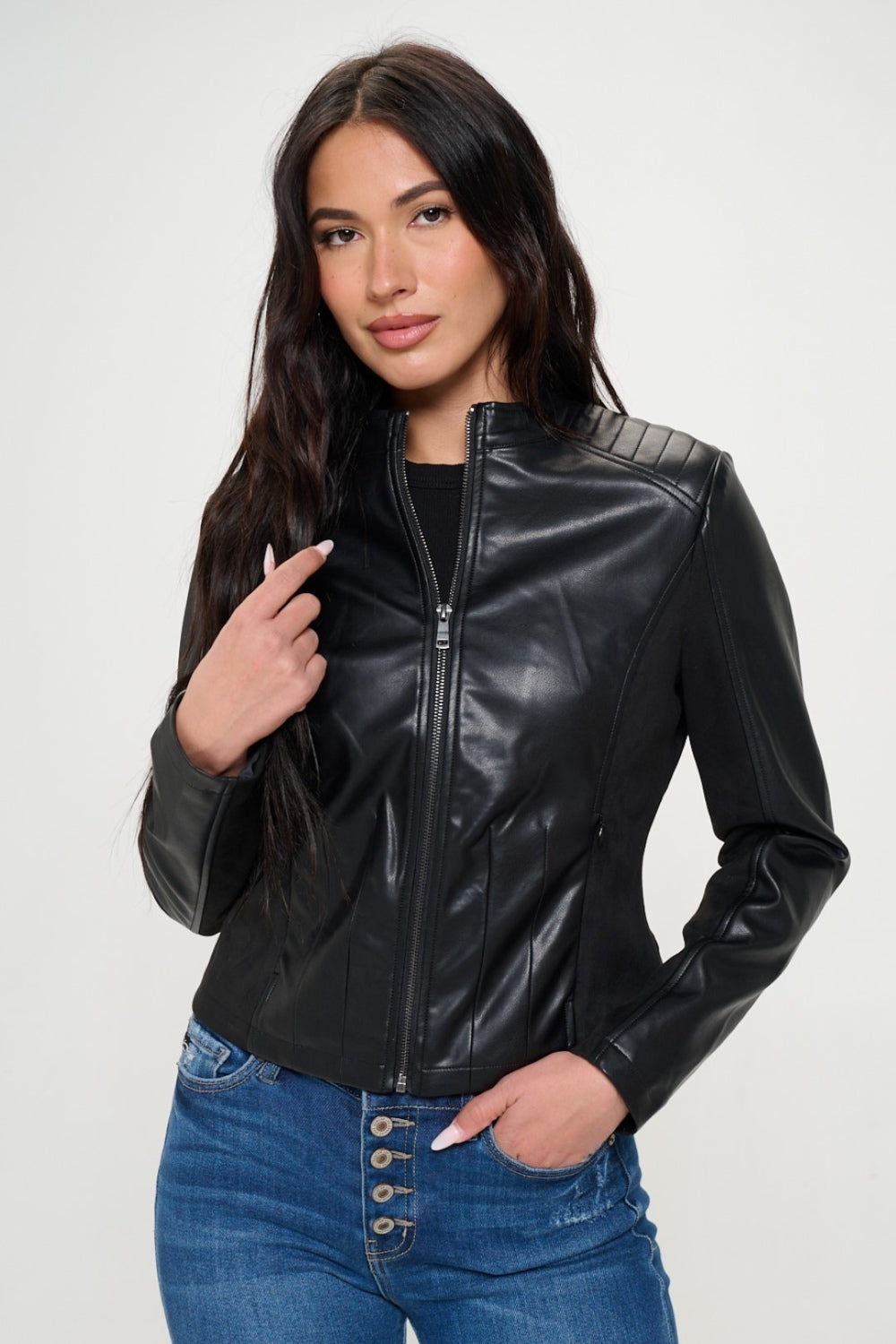 A person with long hair wearing a black Coalition LA Zip Up Vegan Moto Jacket and blue jeans stands against a plain background, showcasing a stylish spring wardrobe.