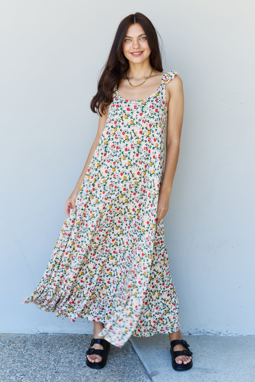 A woman is standing against a gray wall, wearing the Doublju In The Garden Ruffle Floral Maxi Dress in Natural Rose and black sandals, with her left hand raised to her head.