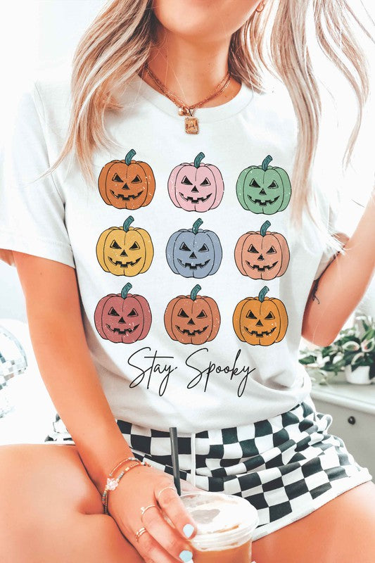 A person wears the STAY SPOOKY MULTI PUMPKINS Graphic Tee in unisex sizing, showcasing nine colorful jack-o'-lanterns and "Stay Spooky" text in a white hue, while holding a drink. They also sport checkered shorts.