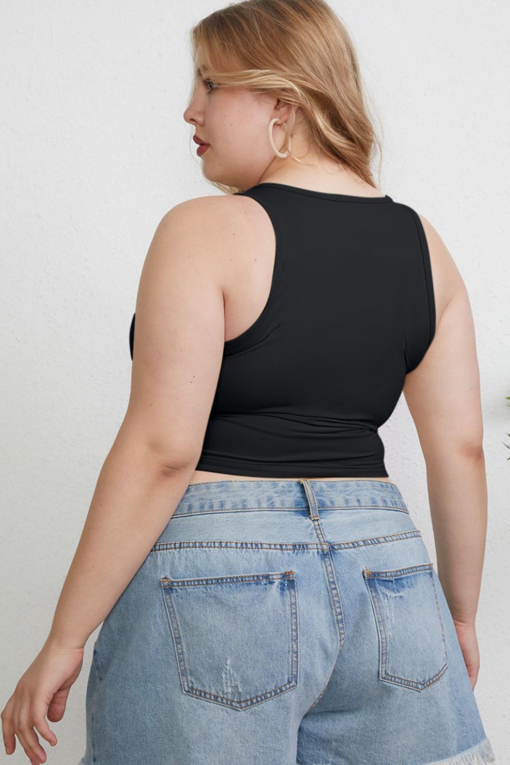 A woman with light hair wears the Basic Bae Full Size Round Neck Slim Tank in black and blue jeans, standing against a light-colored curtain.