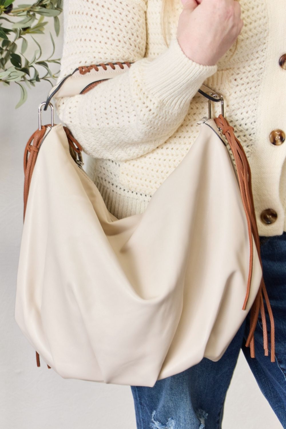 Individual holding a SHOMICO Fringe Detail Contrast Handbag, featuring cream and brown accents, paired with a knitted cream sweater, standing against a light background. The handbag exudes Bohemian flair with its relaxed style.