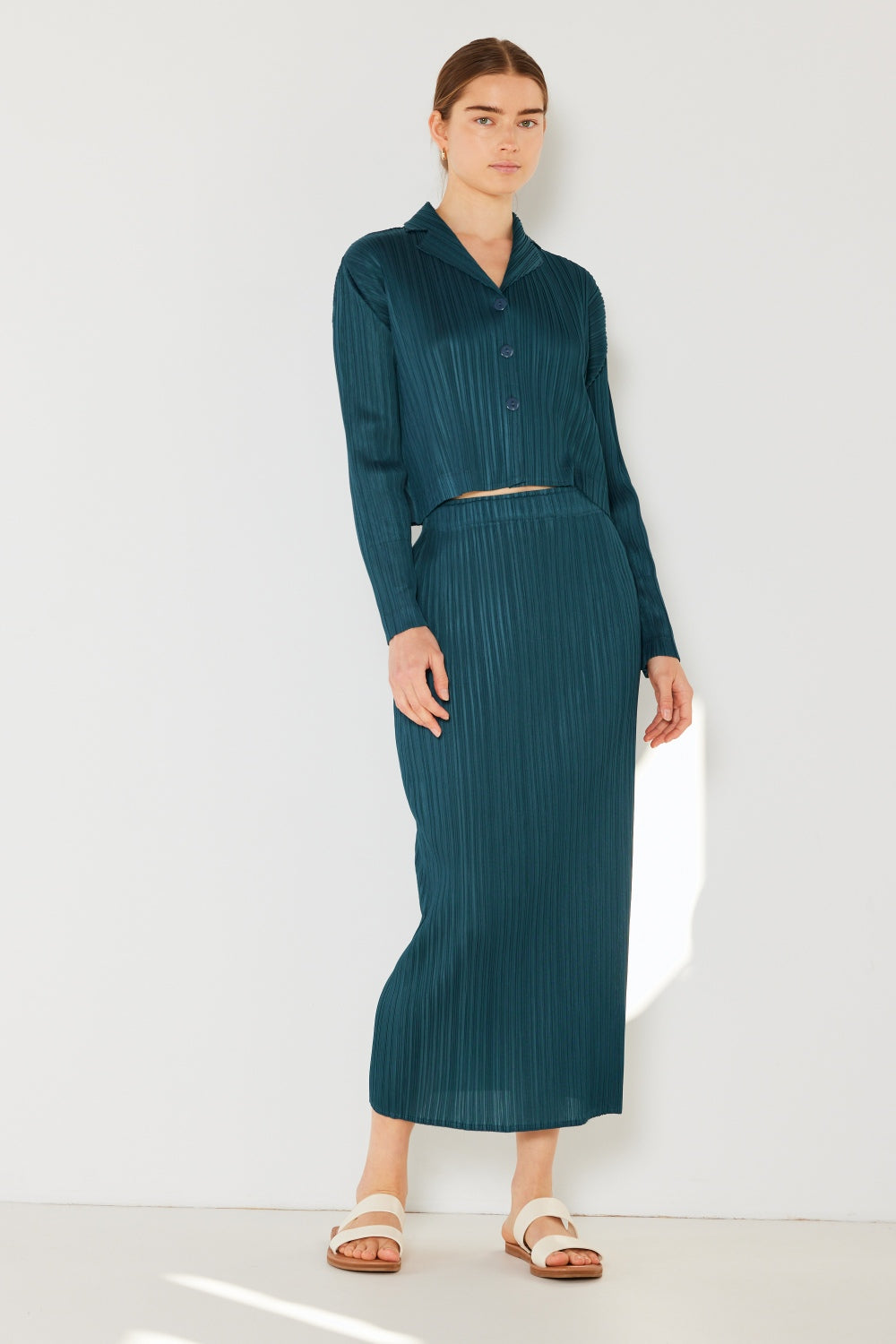 A person wearing the Marina West Swim Pleated Midi Pencil Skirt in teal, paired with a long-sleeve top, stands against a white wall, embodying a classic polished look.