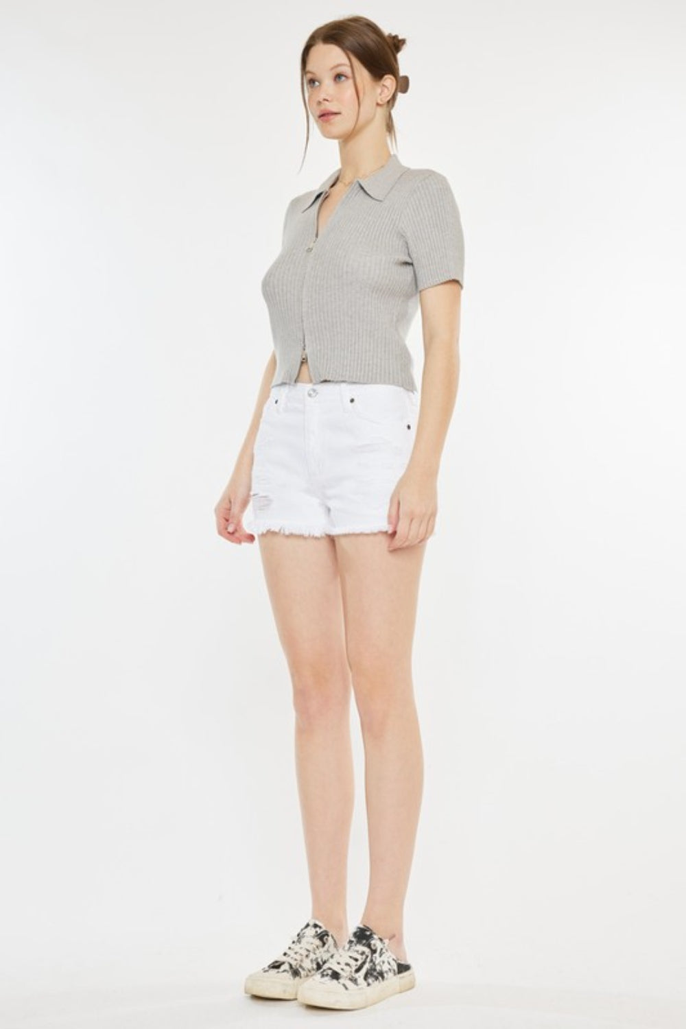 A person stands against a plain background, wearing a light grey short-sleeve top, Kancan Raw Hem Distressed Denim Shorts in white, and white slip-on shoes with black patterns.