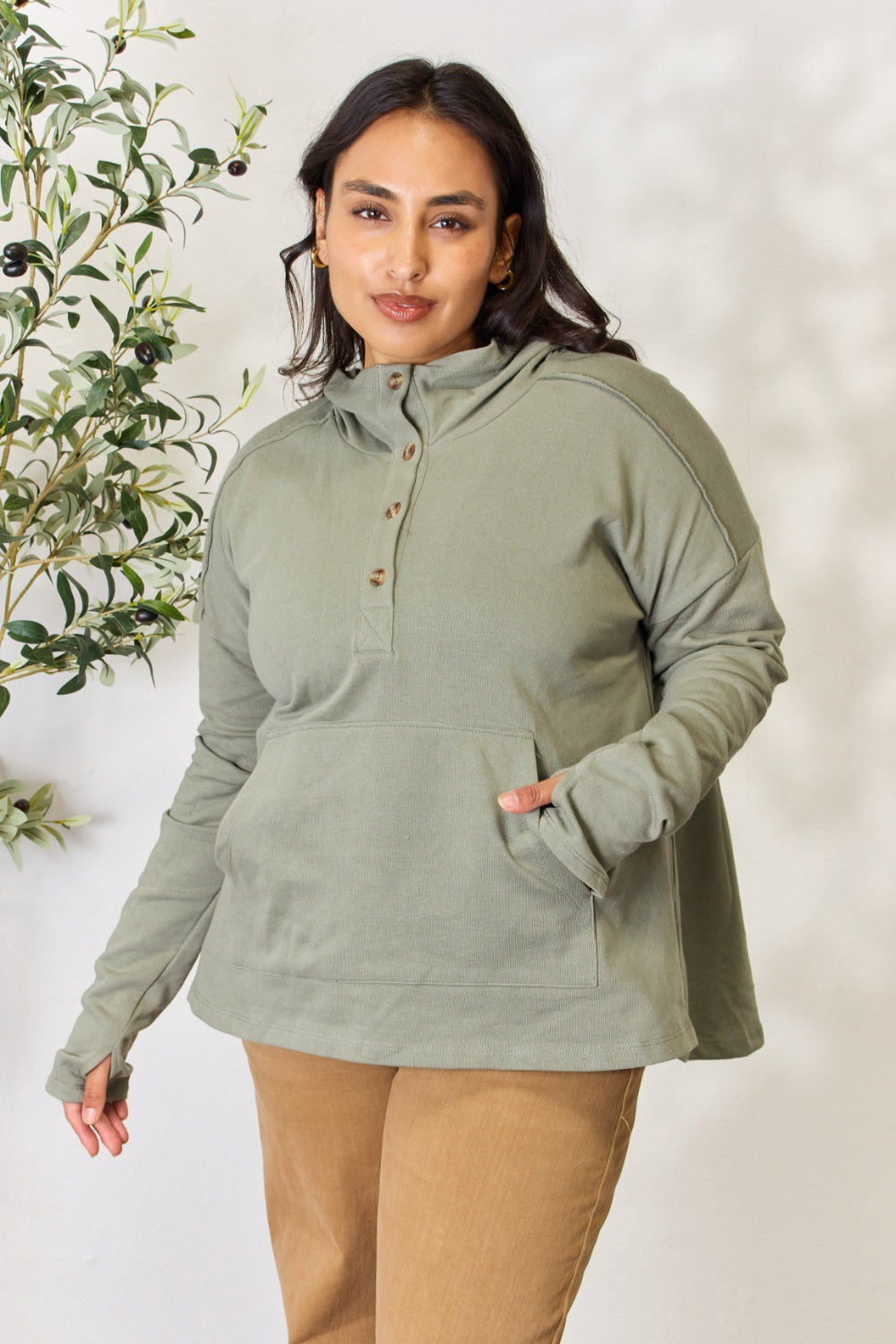 A person stands with hands in the pockets of their light green Culture Code Full Size Half Button Hoodie and brown pants. A plant is visible on the left side of the image.