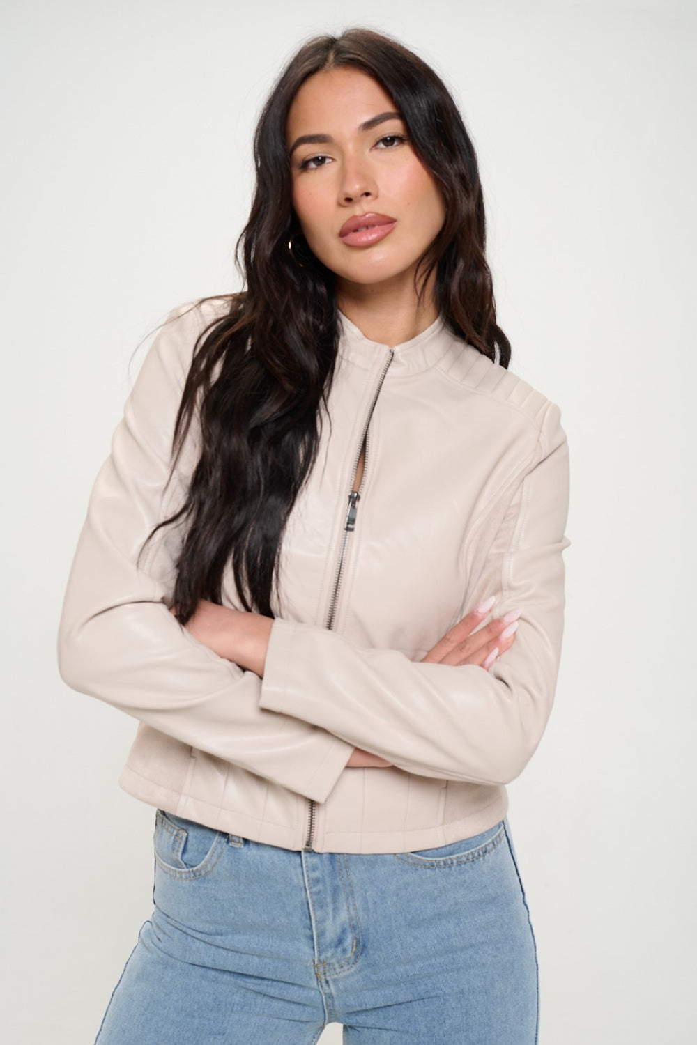 A woman with long dark hair, wearing the cruelty-free Coalition LA Zip Up Vegan Moto Jacket in light beige, pairs it with blue jeans as she stands with her arms crossed.