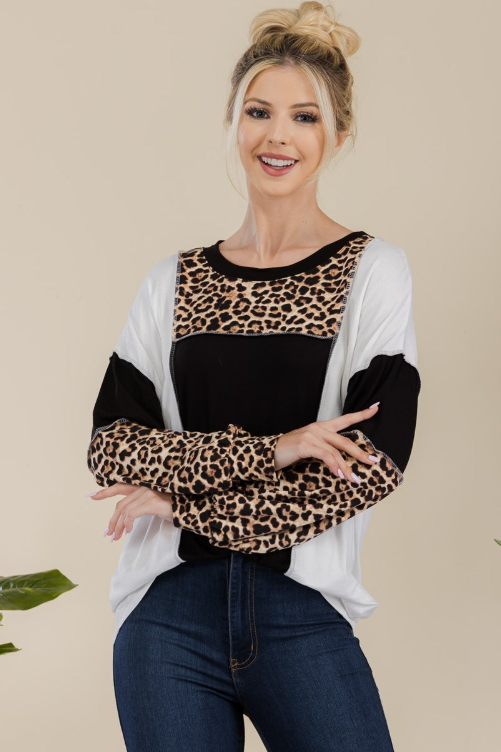 A woman stands in front of a simple background with a plant, wearing the Celeste Full Size Leopard Color Block Exposed Seam T-Shirt, which boasts leopard print cuffs and striking black and white color blocks.