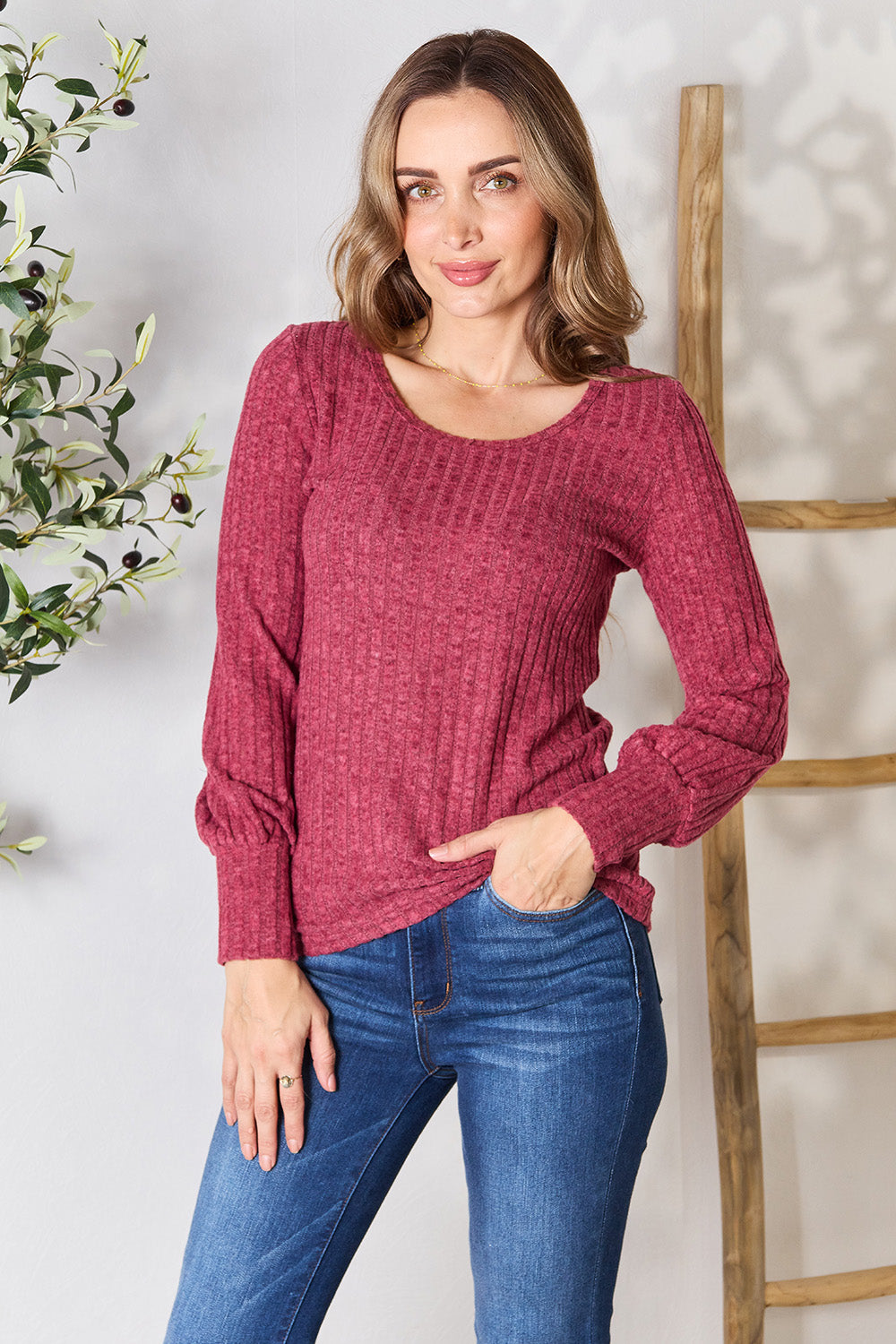 A person in a stylish maroon Ribbed Round Neck Lantern Sleeve Blouse and jeans stands by a wooden ladder, with olive branches in the background.