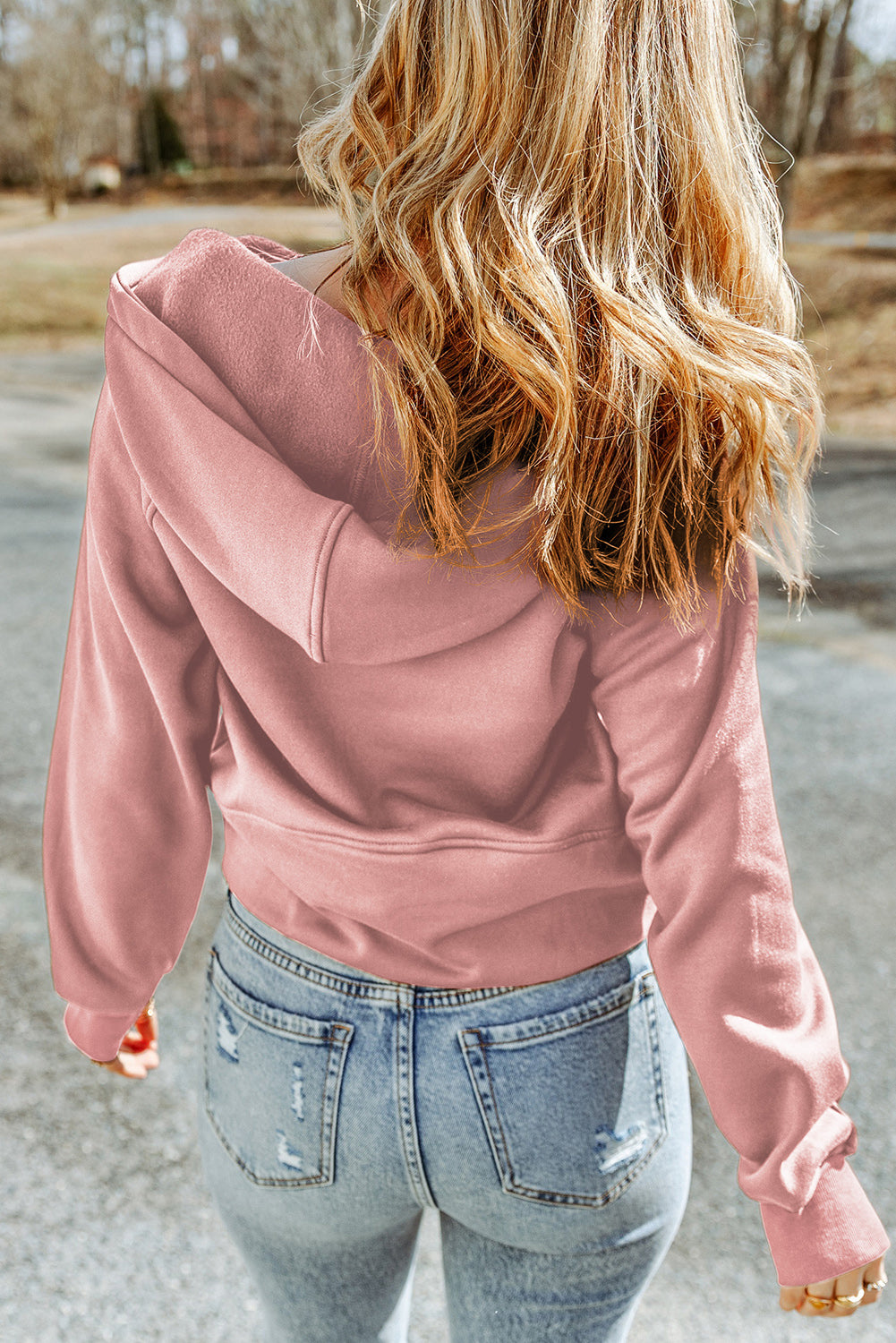 A person with wavy blonde hair is walking outside, sporting a Pink Quarter Zip Kangaroo Pocket Hoodie and light blue distressed jeans. The background includes a road and trees.