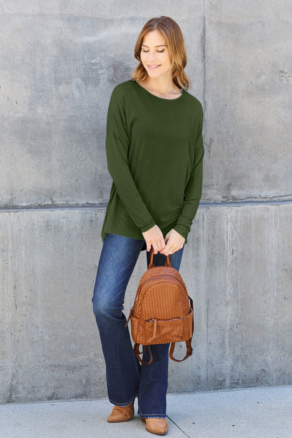 A woman with shoulder-length hair wearing a Basic Bae Full Size Round Neck Dropped Shoulder T-Shirt in green and a pair of jeans, exuding a basic style, stands against a concrete wall. She's holding an imported brown woven backpack on her shoulder while touching her hair with her hand.