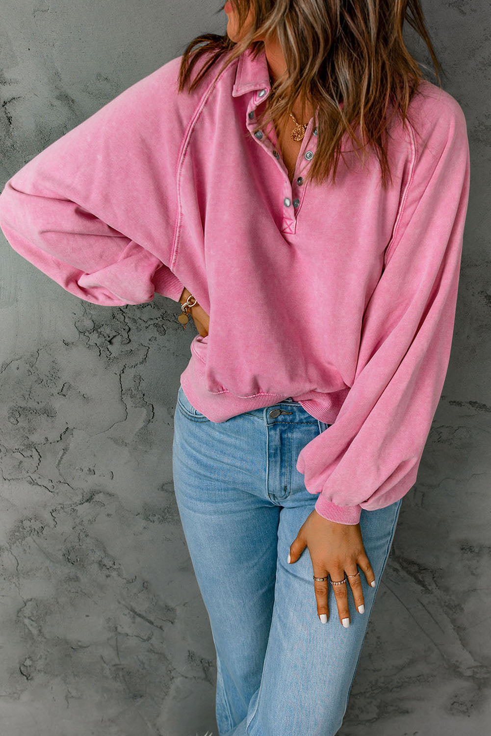 A person with wavy brown hair, wearing a loose-fit Pink Washed Snap Buttons Lantern Sleeve Pullover Sweatshirt and light blue jeans, is standing with their back facing the camera.