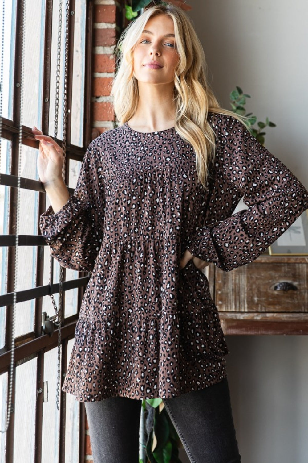 A woman wearing a Heimish Leopard Long Sleeve Tiered Blouse and black pants stands by a window indoors, looking at the camera—embodying a chic and feminine wardrobe.