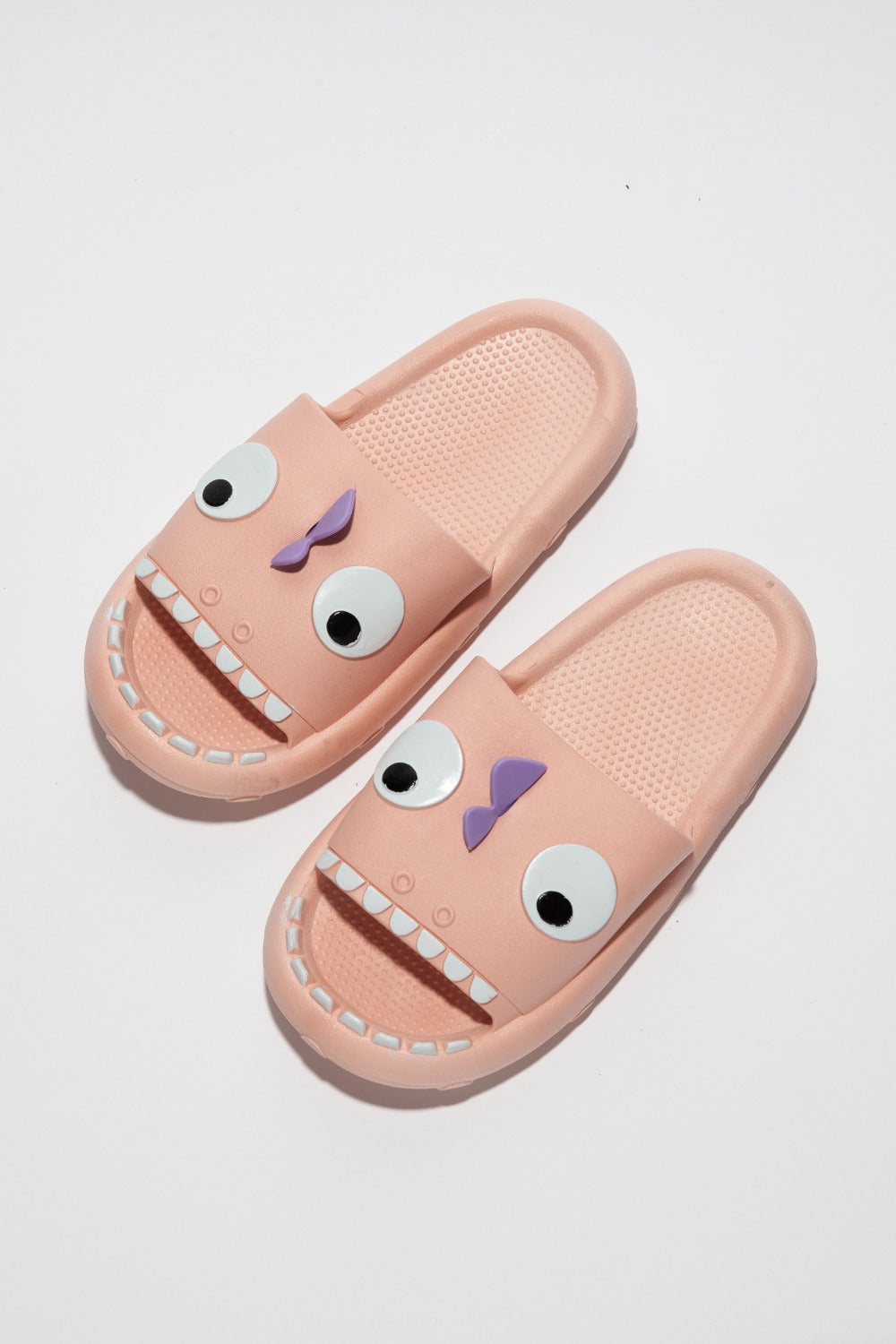 The NOOK JOI Monster Pillow Cloud Slides Non-Slip Slipper features cartoonish monster faces with large eyes, a purple nose, and an open mouth with teeth. Made of durable EVA material, these fun pink cloud slippers also boast an anti-slip design for added safety.