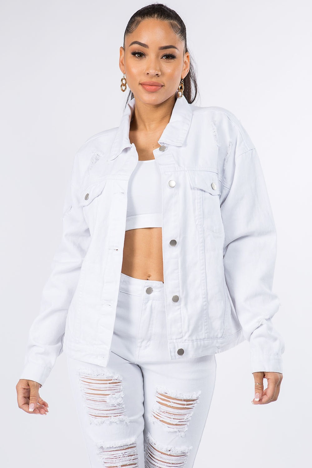 A woman wearing the American Bazi Graphic Distressed Long Sleeve Denim Jacket and matching jeans stands against a plain backdrop. She has her hair styled in a high ponytail and is accessorized with hoop earrings.