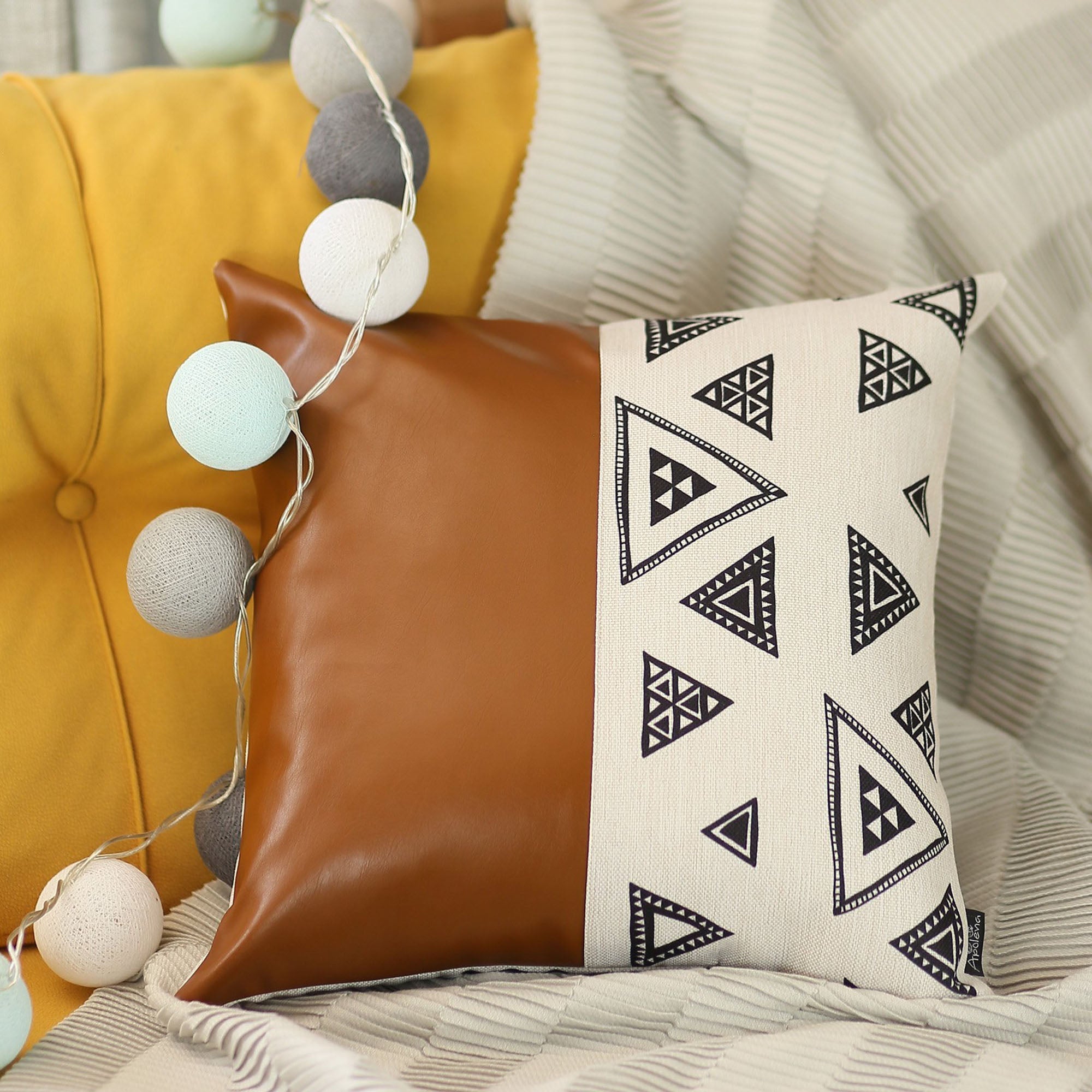 The Bohemian Handmade Decorative Single Throw Pillow, featuring a vegan faux leather geometric square design, rests elegantly on a striped fabric. Nearby, a string of spherical lights illuminates the yellow surface, enhancing the bohemian charm.