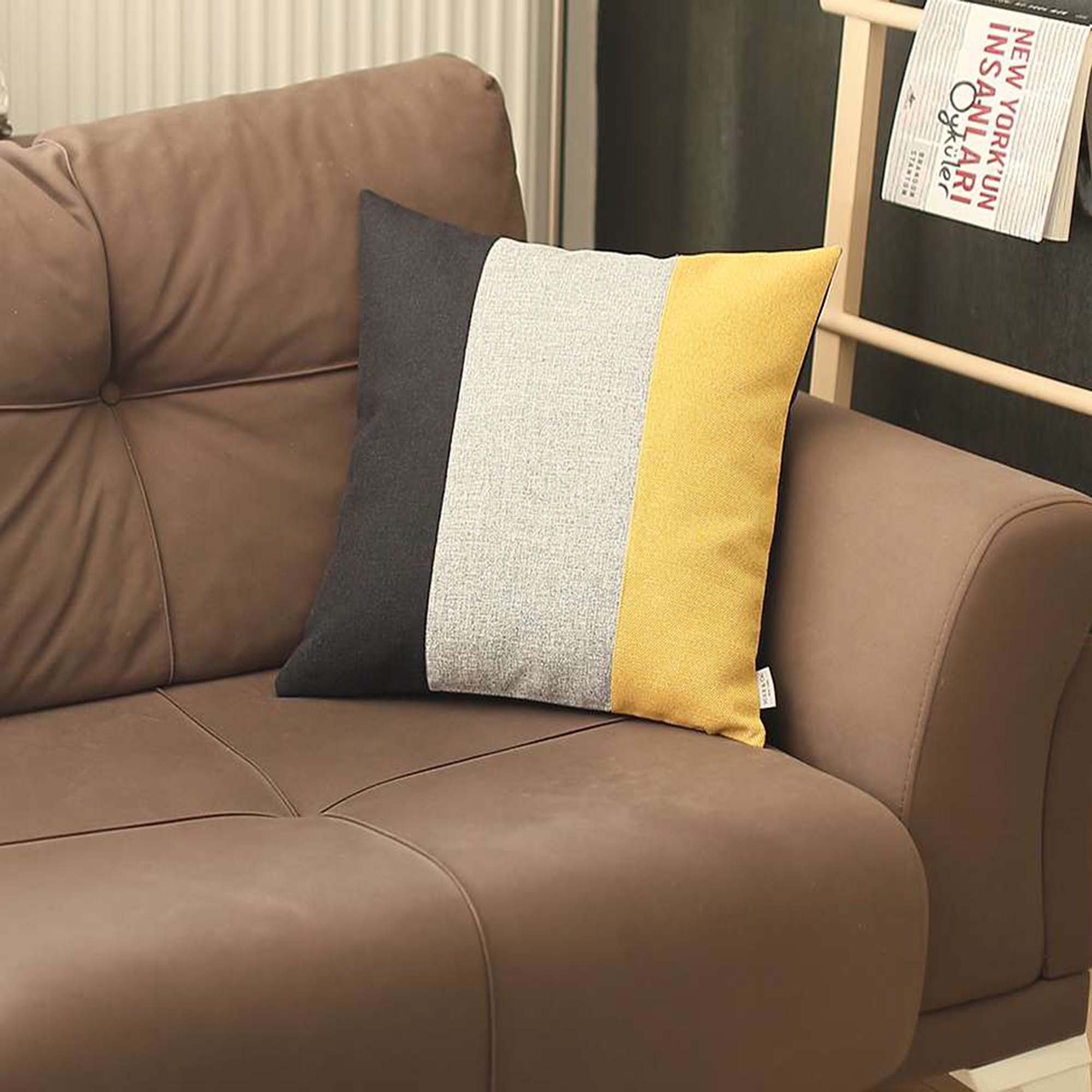 A Bohemian Handmade Decorative Throw Pillow in solid Jacquard, featuring black, gray, and yellow stripes, rests on a brown sofa beside a wooden shelf holding a rolled-up magazine.