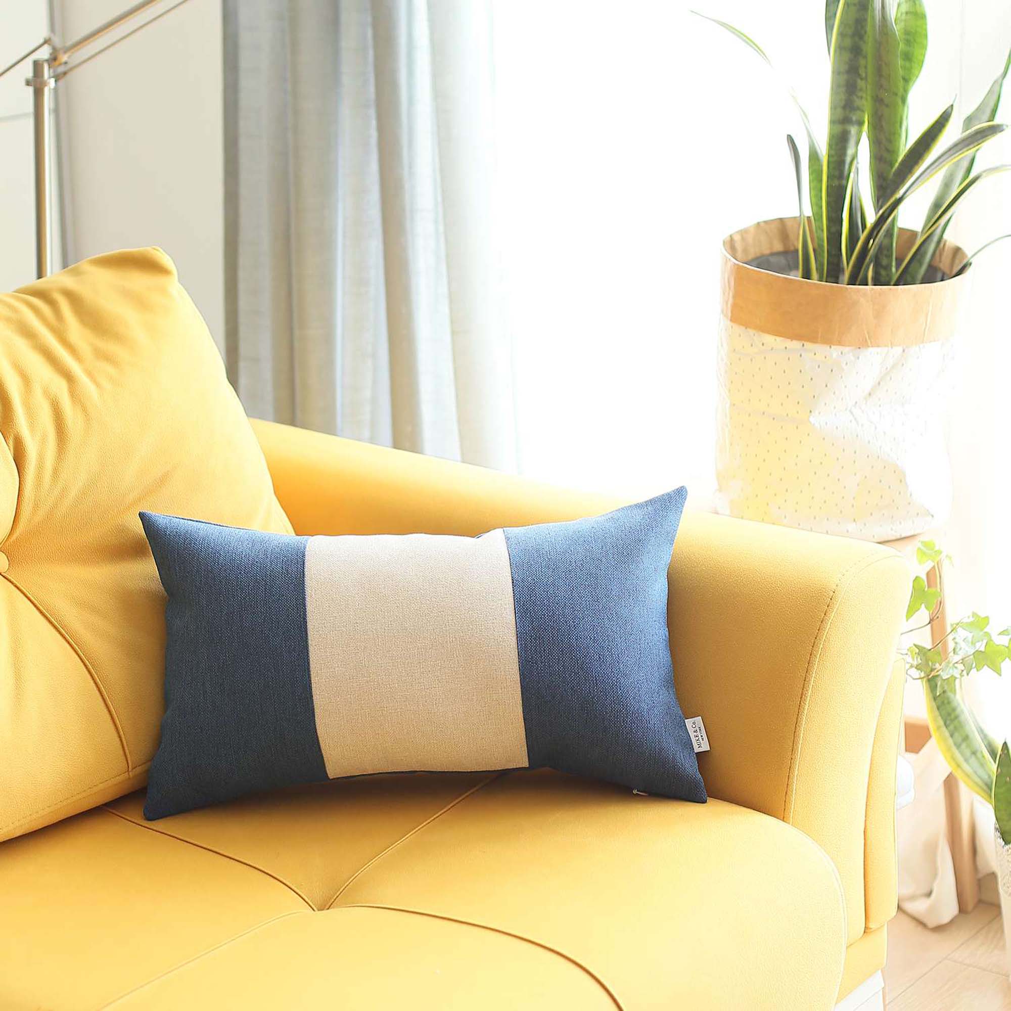 A Bohemian Handmade Decorative Throw Pillow in solid Jacquard, featuring black, gray, and yellow stripes, rests on a brown sofa beside a wooden shelf holding a rolled-up magazine.