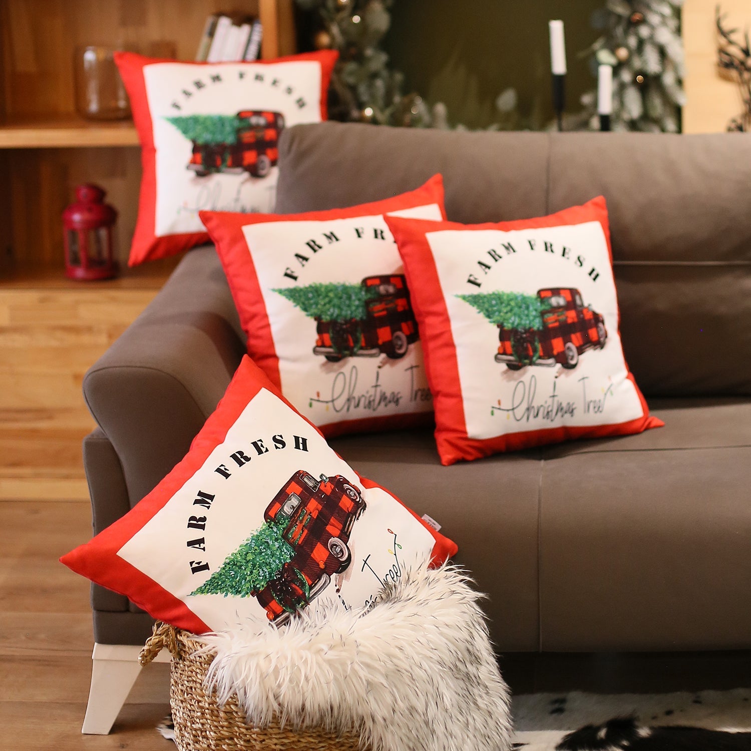 The Christmas Truck Decorative Throw Pillow Set of 4, each measuring 18" x 18" in festive red and white, features a charming design of a red truck carrying a Christmas tree with the "Farm Fresh" text. These pillows are perfect for adding holiday warmth to your sofa or bedding. The cozy setup is enhanced by a basket and fur blanket on the floor, while other festive decor brightens the background. Conveniently machine washable pillow inserts make this an ideal choice for your holiday decor.