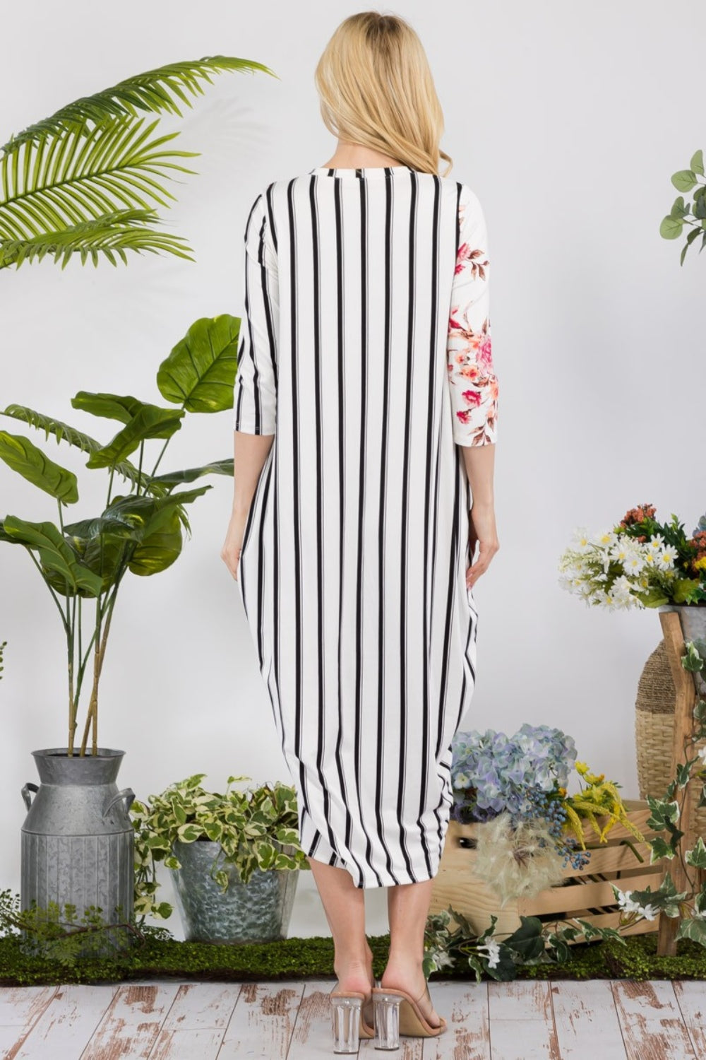 Dressed in the Celeste Full Size Floral Striped Contrast Midi-Dress with Pockets, a person stands elegantly among potted plants and flowers on a wooden floor, highlighting the dress's versatile styling options.