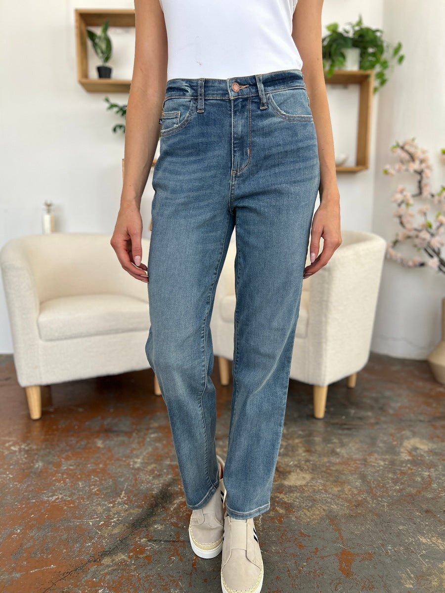 Person wearing Judy Blue Full Size High Waist Straight Jeans, a white shirt, and black sandals stands on a polished concrete floor with a white armchair in the background.