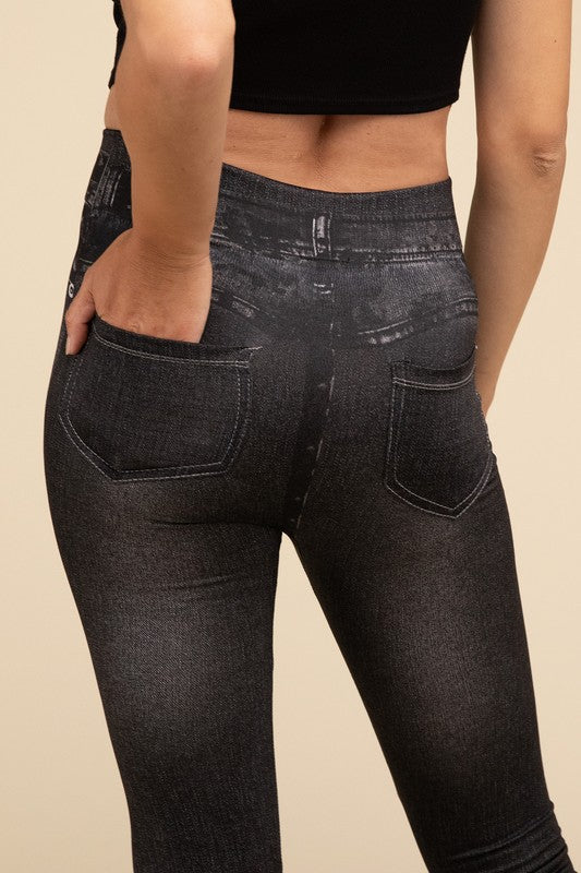 A person wearing Denim Leggings in a high-waisted black design and paired with black heels poses against a plain beige background, highlighting the sleek, stretchy fit.