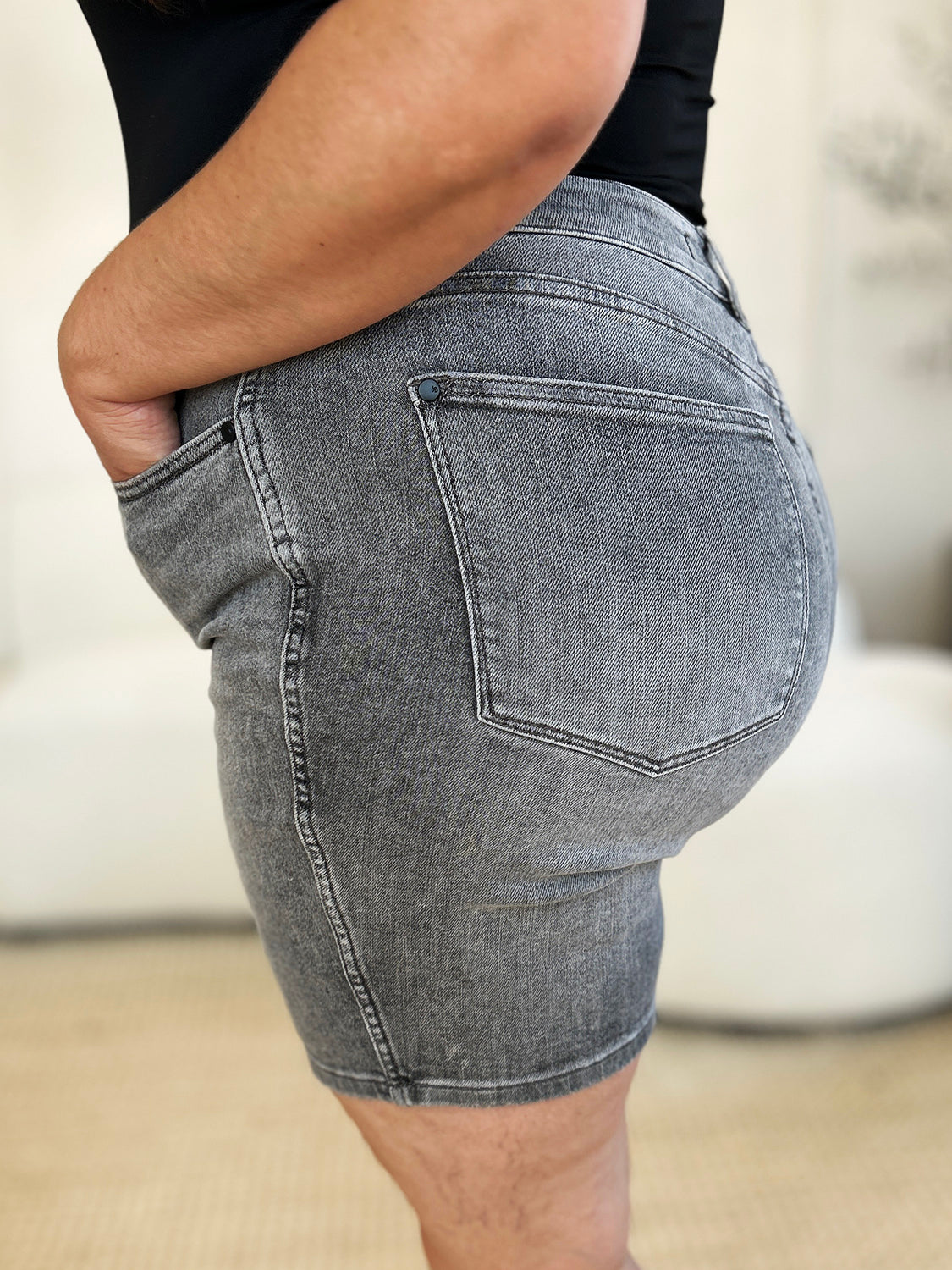 A person wearing a white top and the Judy Blue Full Size High Waist Washed Denim Shorts with vintage charm has one hand in their pocket. The shorts, featuring a faded design and belt loops, contribute to a retro-inspired look.