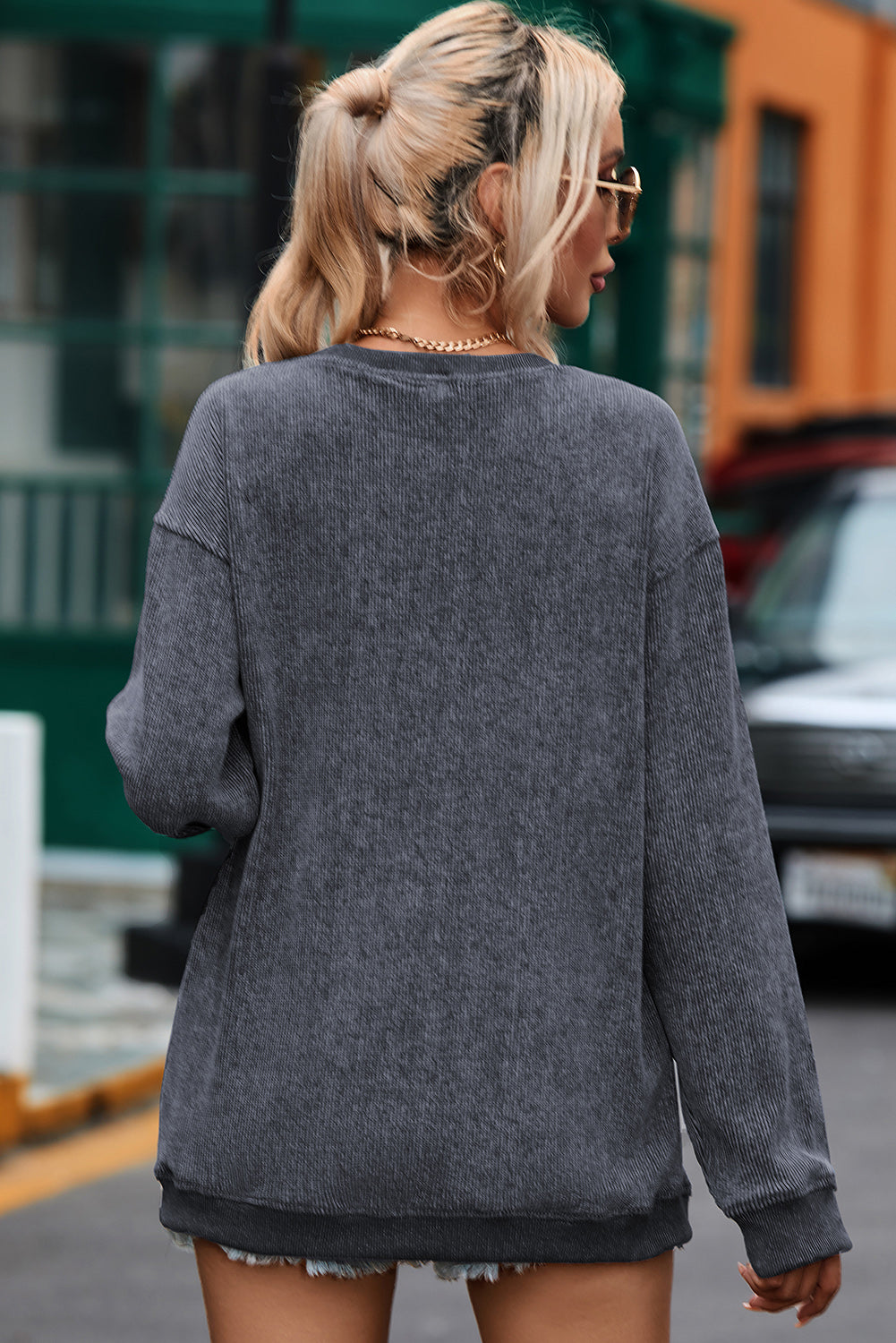 A person with long, blonde hair smiles while wearing an oversized Gray Solid Ribbed Knit Round Neck Pullover Sweatshirt and a gold chain necklace. They are standing indoors near a window, embodying versatile style.