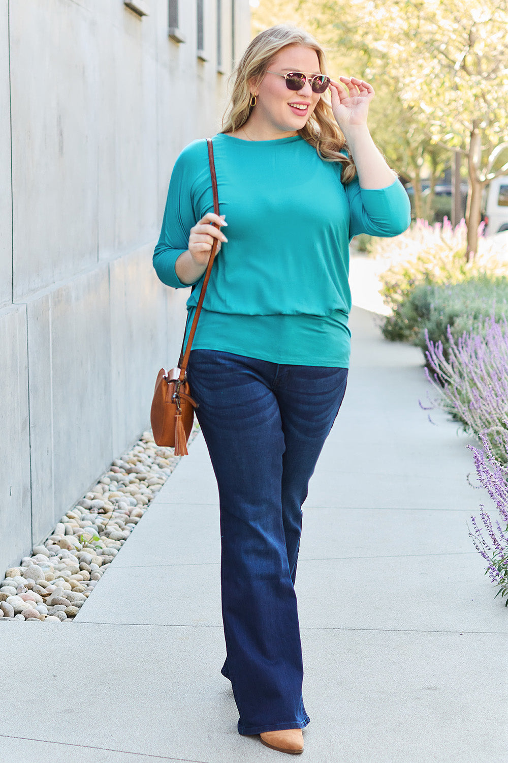 A woman in a slightly stretchy, brown Basic Bae Full Size Round Neck Batwing Sleeve top and blue jeans stands against a concrete wall, holding a brown handbag.