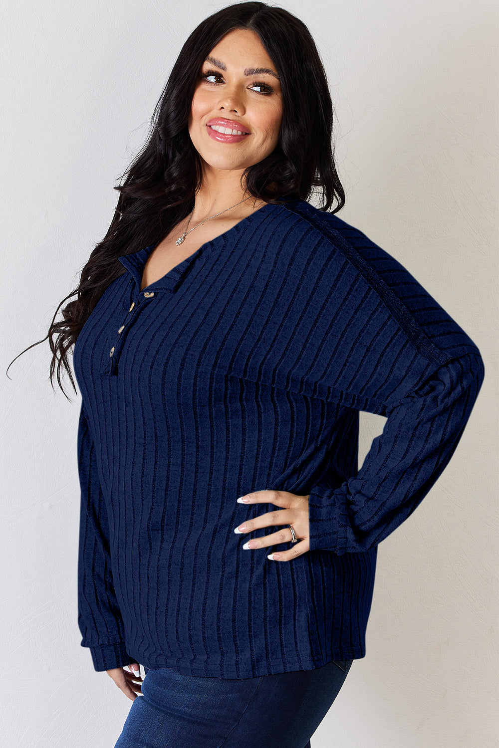 A woman with long, wavy blonde hair wears a slightly stretchy, textured Basic Bae Full Size Ribbed Half Button Long Sleeve T-Shirt in pink, standing against a neutral backdrop.