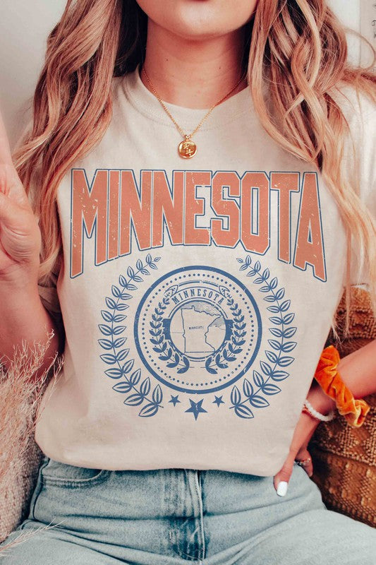 A woman sporting a 100% cotton MINNESOTA Graphic Tee with "Minnesota" emblazoned in bold orange letters and a decorative state emblem underneath, complemented by high-waisted denim shorts and a plaid shirt casually draped over her shoulders. The tee offers unisex sizing for an extra comfortable fit.