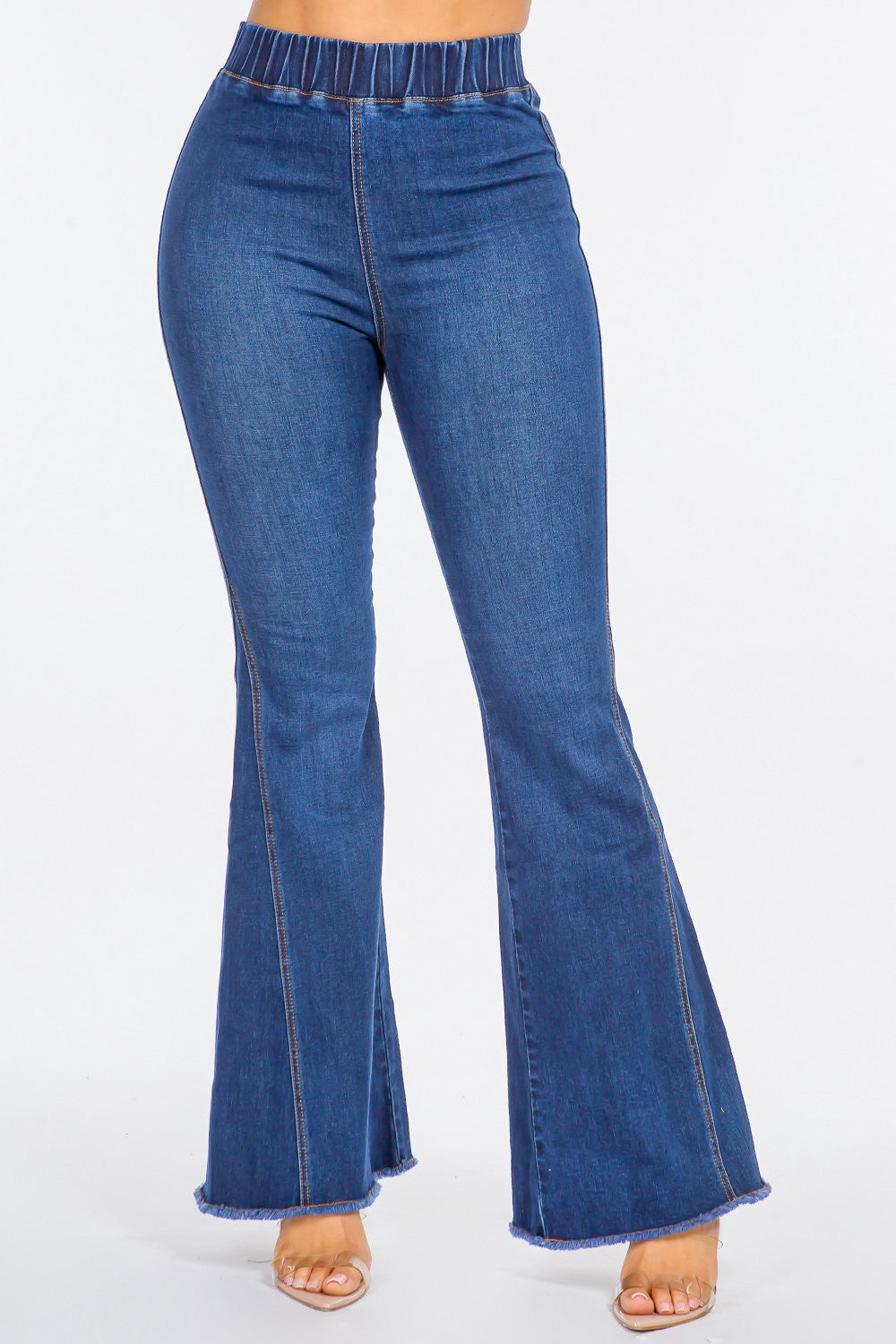 The image shows a person wearing American Bazi High Waist Curvy Flare Jeans made from stretchy denim, paired with nude open-toe heels. The jeans feature an elastic waistband and frayed hems, adding a touch of retro flair. The background is plain white.