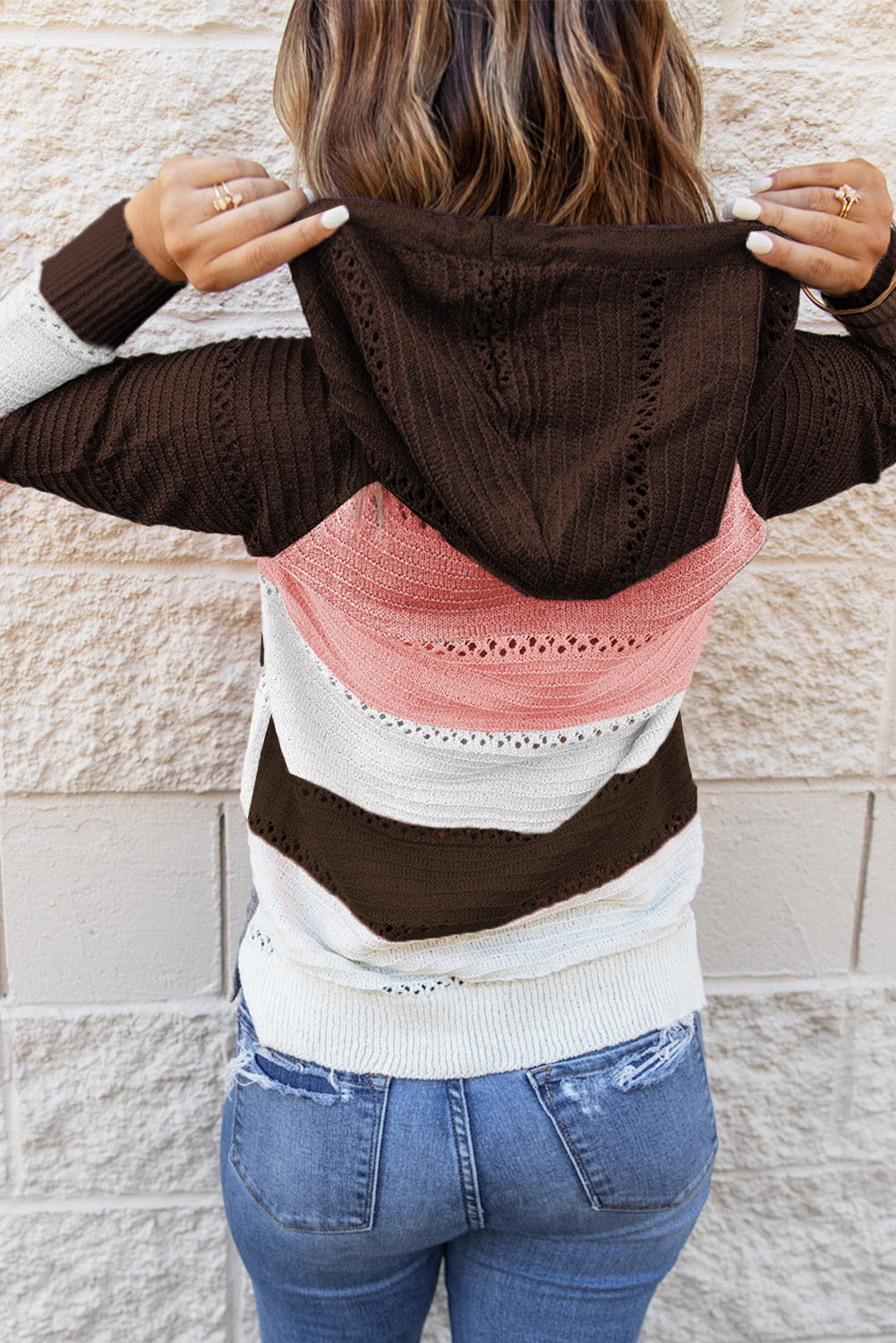 A person stands with their back to the camera, adjusting the hood of a Brown Beach Bonfire Knitted Hoodie featuring dark brown, pink, white, and light brown stripes. Perfect for a casual style, they are wearing distressed blue jeans and showcasing the laid-back vibe of lightweight hoodies.