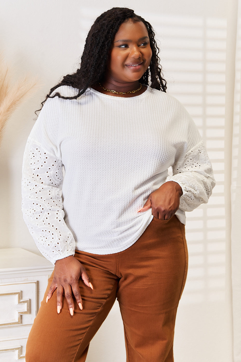 A woman wearing a Shiny Eyelet Dropped Shoulder Round Neck Blouse and brown pants stands in a room with sunlight streaming through blinds. She is smiling and has her left hand on her hip.