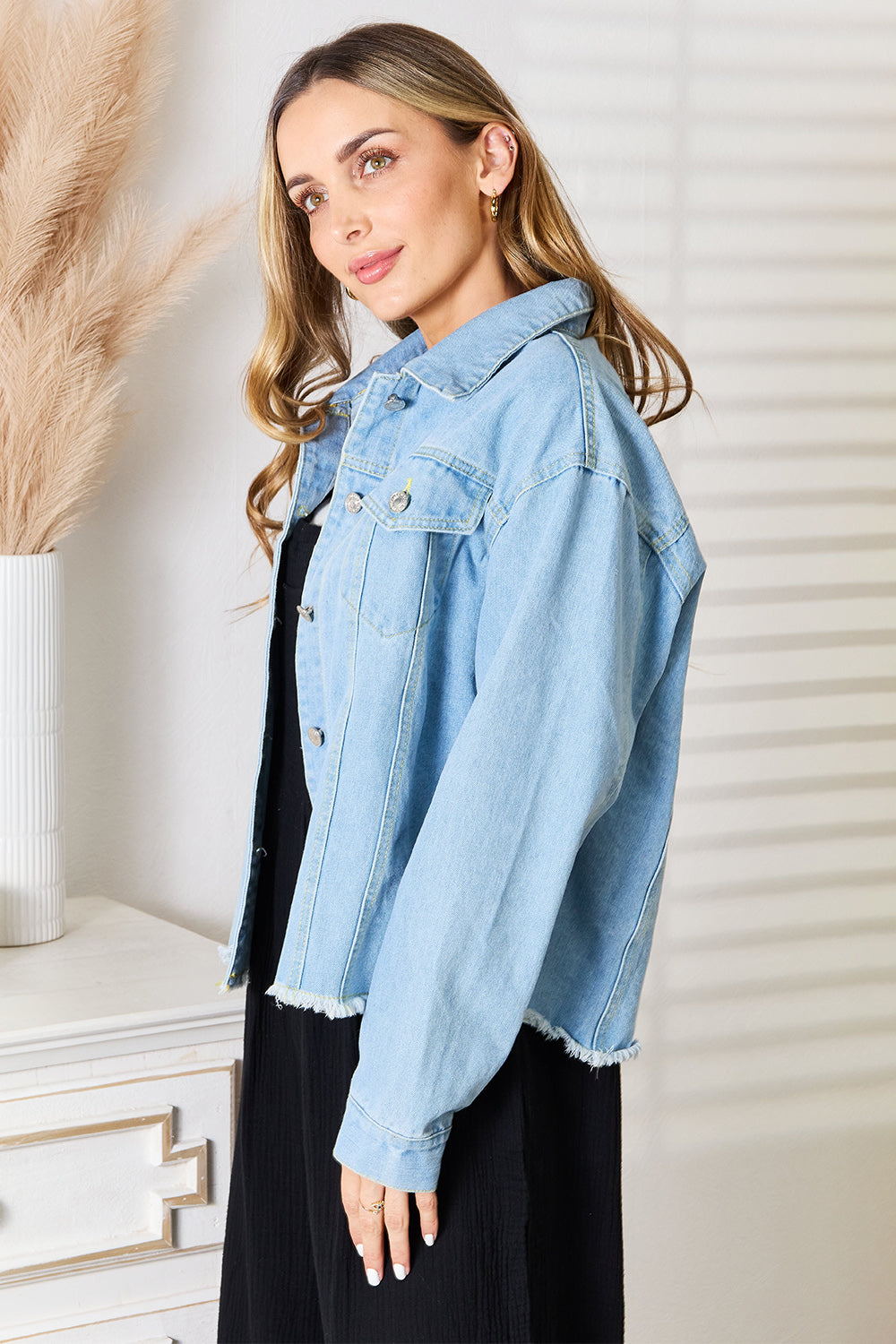 A woman with long hair wearing the Double Take Dropped Shoulder Raw Hem Denim Jacket over a black dress stands indoors near a white cabinet adorned with decorative plants.