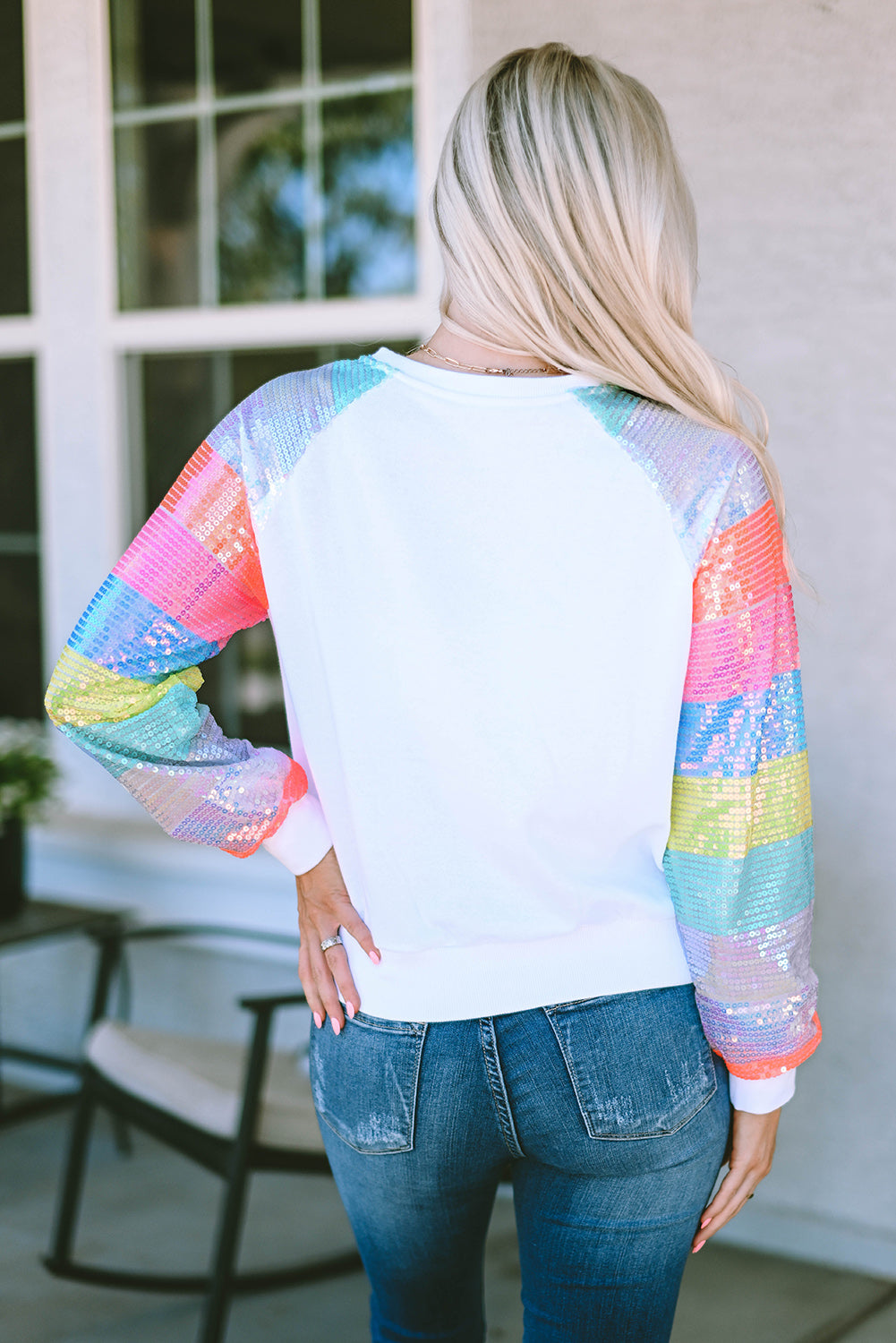 A person with long blonde hair is seen from the back, wearing a White Sequin Color Block Raglan Sleeve Pullover Sweatshirt and blue jeans. They are standing in front of a building with large windows.