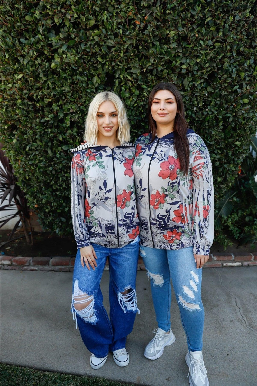 A woman stands outdoors wearing the Blumin Apparel Full Size Floral Zip-Up Hoodie with her hands in the pockets. A leafy green hedge is in the background, adding to the casual chic vibe.