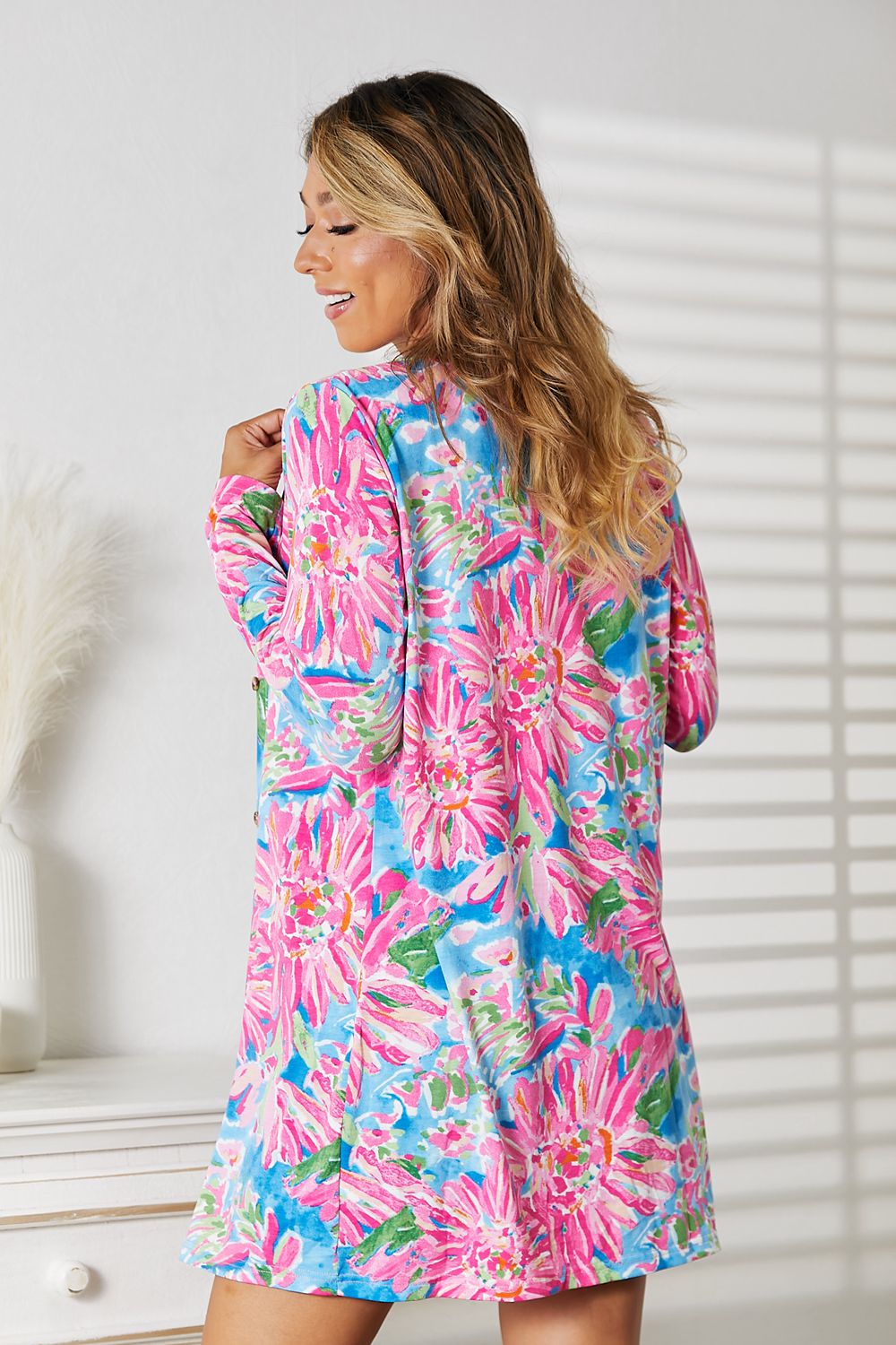 Wearing the Double Take Floral Open Front Long Sleeve Cardigan in shades of pink and blue over a white top and denim shorts, a person exudes feminine charm while standing in a well-lit room, smiling.