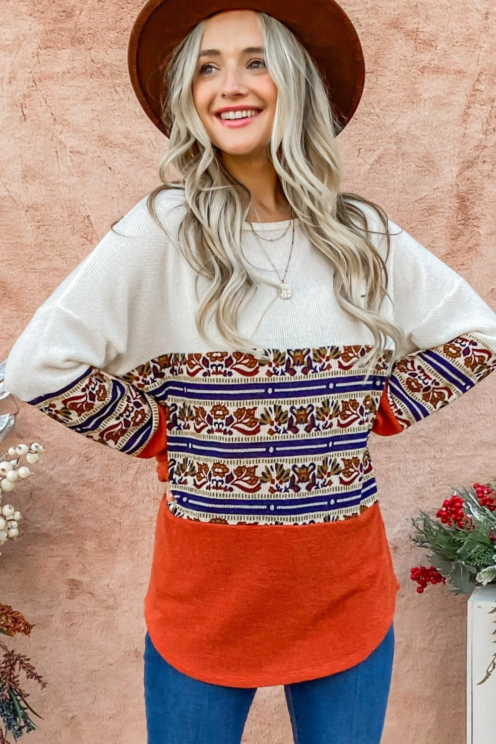 A woman with long blonde hair wearing a brown hat and the And The Why Ethnic Print Color Block Top stands against a peach wall next to a floral arrangement.