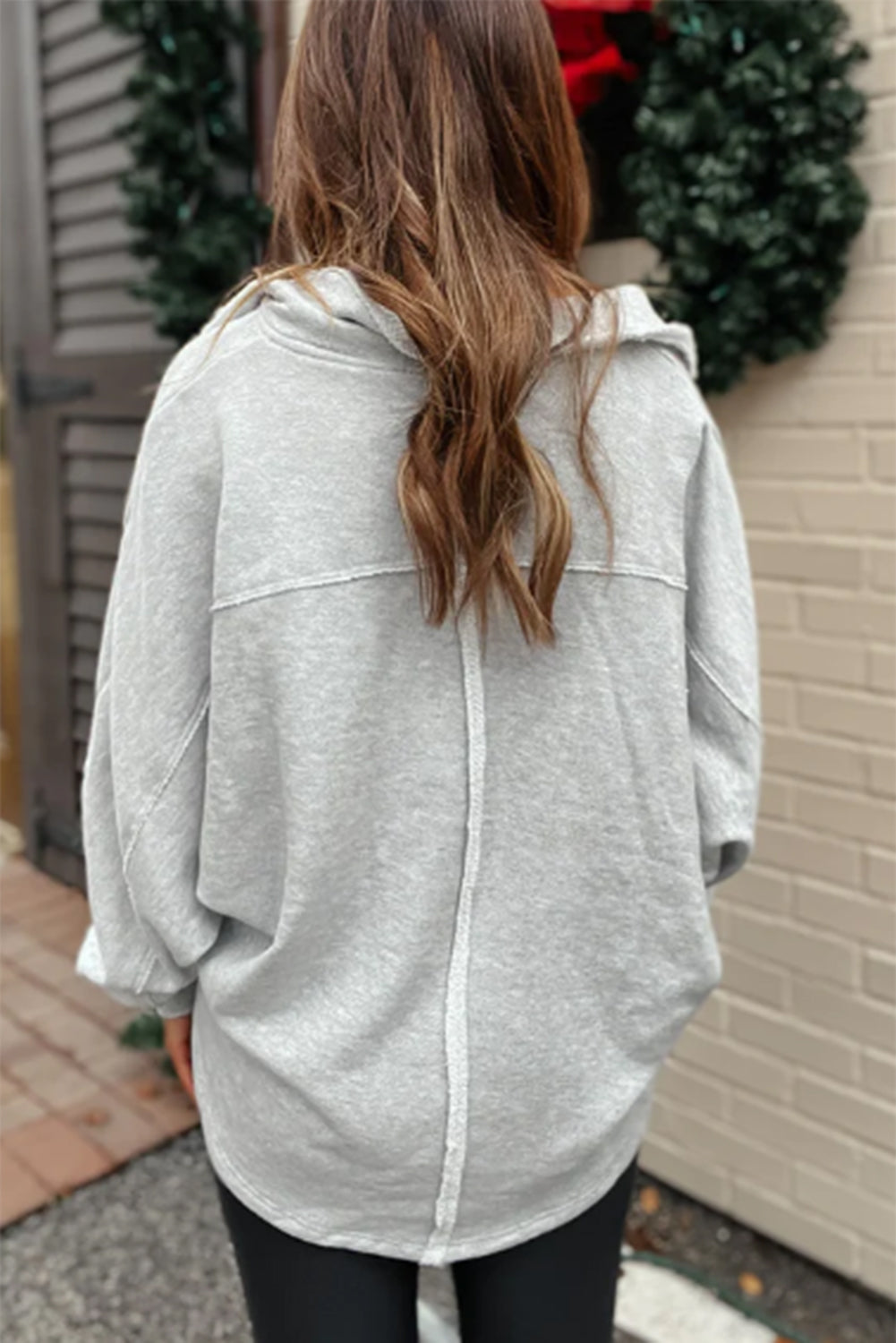 A person with long brown hair is seen from the back wearing a loose Gray Exposed Seam Double Chest Pocket Sweatshirt and black pants, standing outside near a building adorned with wreaths.